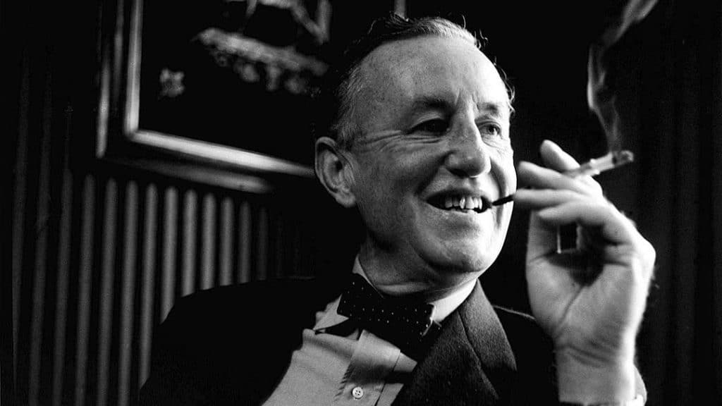 Black-and-white photo of Ian Fleming smiling while holding a cigarette.
