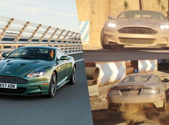 Aston Martin DBS in silver from 007 First Light Story Trailer driving through a Mediterranean harbor area, compared with a green production DBS on an open road.